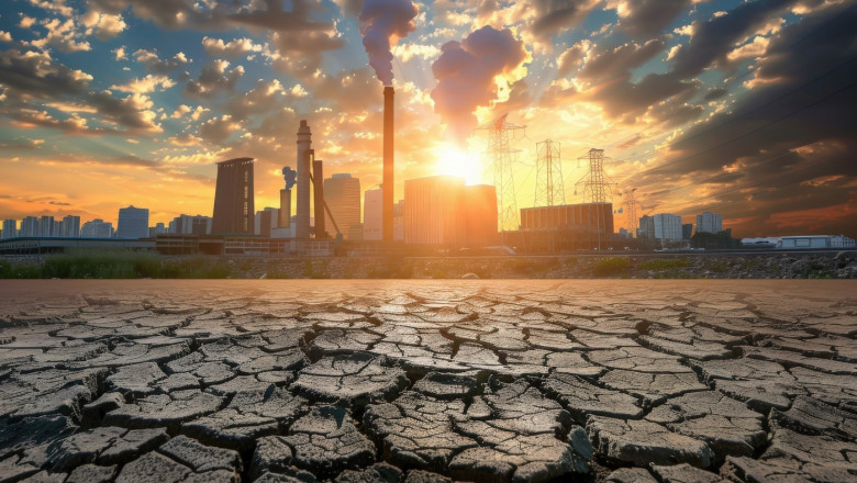 A factory spews smoke into the air as the sun sets over a cracked, dry landscape.