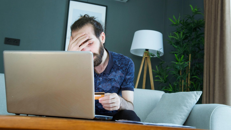 A man sits on a couch, distressed, holding his forehead with one hand and a credit card in the other. Frustrated and worried, highlighting financial s