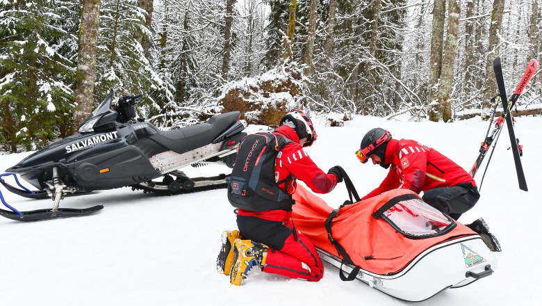 2 salvamontisti cu o targa-sanie, snowmobil in spatele lor