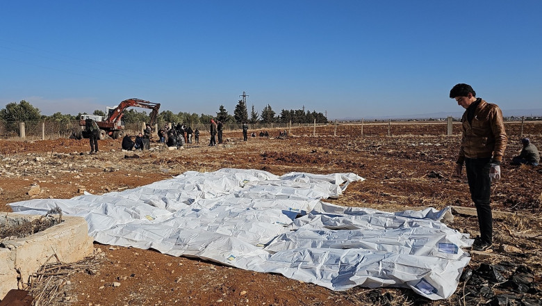 cadavre in saci, un om se uita, excavatior in fundal - groapa comuna descoperita in siria