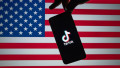 Stone / United Kingdom - July 22 2020: TikTok logo seen on the silhouette of smartphone hold in a hand with blurred American flag on tha back. Rela p