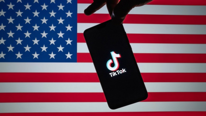 Stone / United Kingdom - July 22 2020: TikTok logo seen on the silhouette of smartphone hold in a hand with blurred American flag on tha back. Rela p