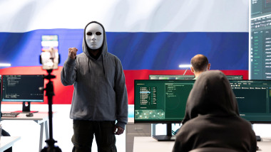 Anonymous person with mask asks for ransom in Russian governmental hacking room with the national flag. Dealing with cyber espionage, disinformation campaigns and hybrid war propaganda.