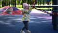 Baby girl playing in children playground in city park in cold weather spring. Toddler girl walking expressively in public playground in sunny day.