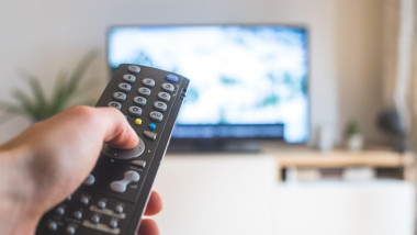 Holding a TV remote control in the hand, foreground, tv in the blurry background. Streaming.