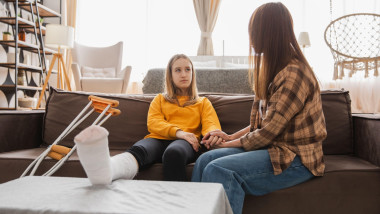 Girl with broken leg in cast sits with mother on couch at home. Sad little kid hugging teddy bear recovering from broken leg. Girl using cast crutches for quick rehabilitation. Mom supporting daughter
