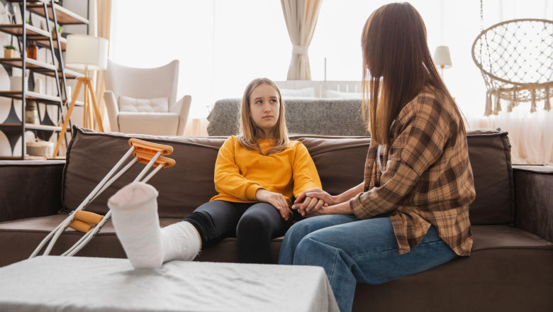 Girl with broken leg in cast sits with mother on couch at home. Sad little kid hugging teddy bear recovering from broken leg. Girl using cast crutches for quick rehabilitation. Mom supporting daughter