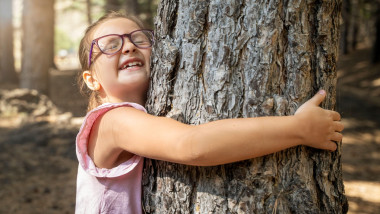 A happy young girl with glasses joyfully closes her eyes and hugs a large tree trunk, showing love and connection with nature in a forest