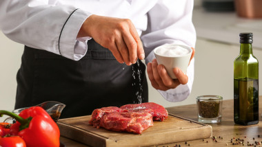 Male chef salting raw meat at table in kitchen, closeup