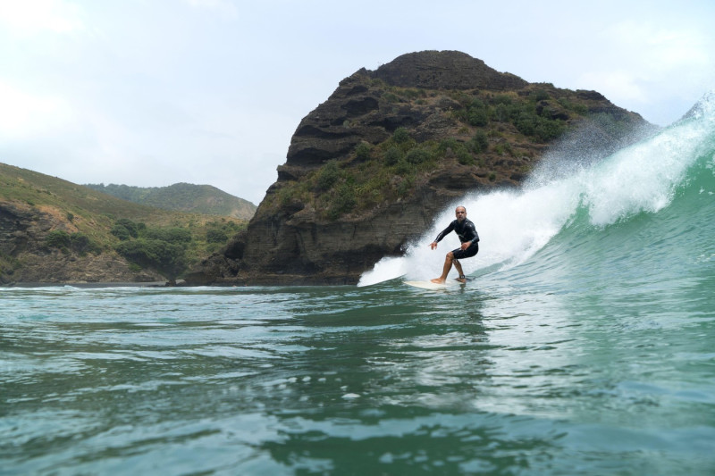 A middle aged man surfs on a large green breaking wave at Piha Beach, Auckland, New Zealand. Rocky coastline in the background, blue sky with clouds.