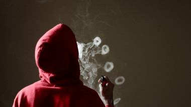 Young male in red hoodie vaping smoking, blows multiple smoke rings while holding a vape in hand, isolated rear view