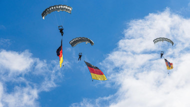 Two German Bundeswehr paratroopers descend under parachutes, proudly carrying the German flag against a clear blue sky, symbolizing strength, unity, a