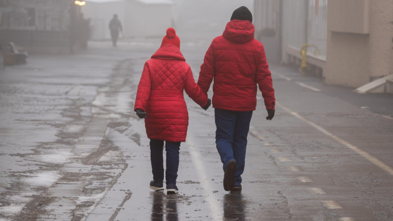 două persoane îmbrăcate în haine groase de iarnă merg prin ceață pe o stradă umedă.