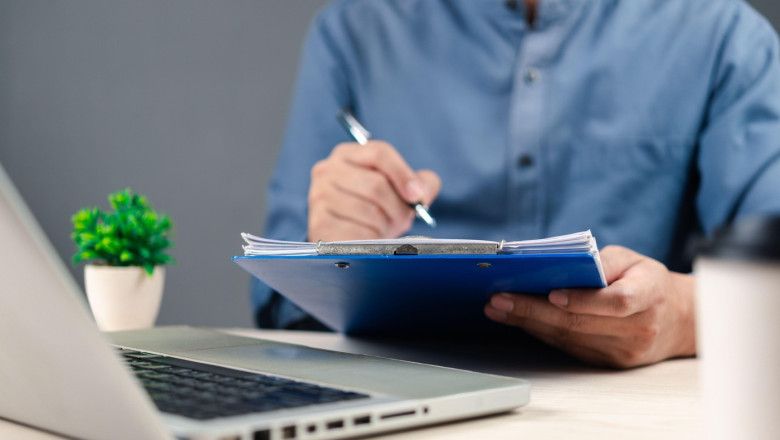 A professional in a blue shirt holding a clipboard and writing, with a laptop and small plant in the background, symbolizing office work, documentatio
