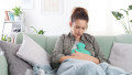Young woman sitting on sofa at home suffering from period pain, cramps. Holding hot water bottle on abdomen, dealing with endometriosis, indigestion,