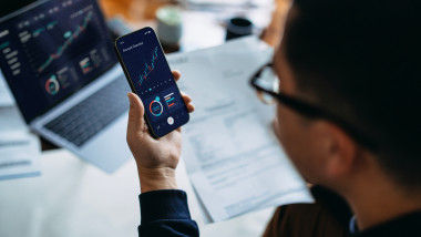 Young Asian man managing finance and investment online, analyzing stock market trades with smartphone and laptop. Making financial plans. Banking and finance, investment, financial trading, mobile banking with technology concept