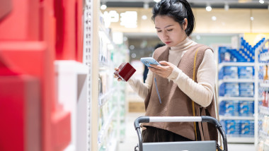 An Asian woman uses her mobile phone to check discount information while selecting cosmetics in a supermarket.