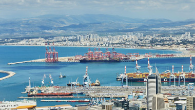 Haifa harbor and city skyline with container cranes, cargo ships, and residential buildings under a cloudy mountain backdrop