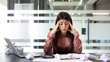 Young businesswoman clutching her head at a cluttered desk with crumpled papers and laptop, overwhelmed by deadlines and burnout, stressed and exhausted at work
