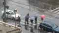 People walk across a busy street while holding umbrellas on a rainy day. Cars are parked nearby, and the wet pavement shines under the falling rain.