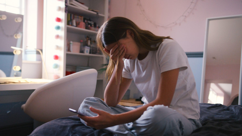 Depressed Teenage Girl Sitting On Bed At Home Looking At Mobile Phone With Head In Hands