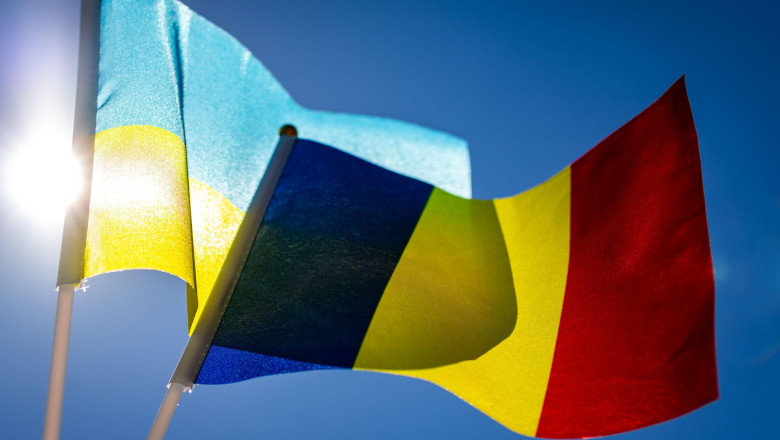 Backlit Ukrainian and Romanian flags billow in a cloudless sky. Sharp color and movement highlight solidarity partnership and the desire for stability