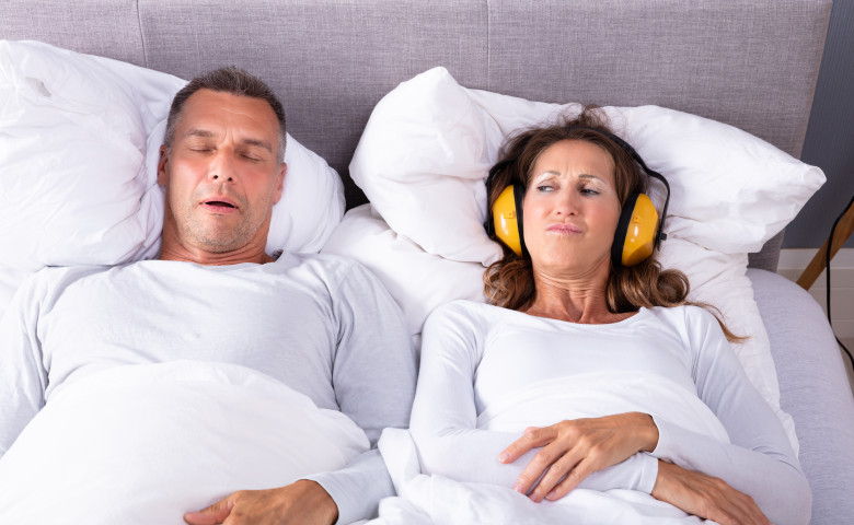 Mature,Woman,Covering,Her,Ears,With,Headphone,While,Man,Snoring