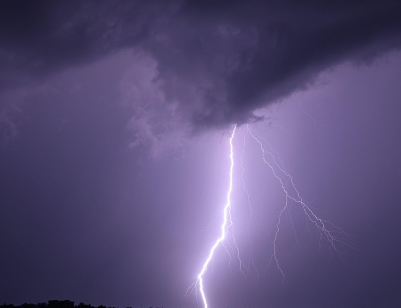 Single powerful lightning bolt hitting the ground from dramatic storm clouds at night, illuminating the dark sky above a city skyline. Long exposure n