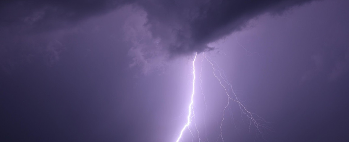 Single powerful lightning bolt hitting the ground from dramatic storm clouds at night, illuminating the dark sky above a city skyline. Long exposure n