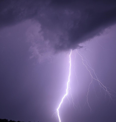 Single powerful lightning bolt hitting the ground from dramatic storm clouds at night, illuminating the dark sky above a city skyline. Long exposure n