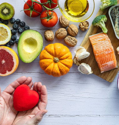 Two hands holding a red heart surrounded by healthy foods to prevent cancer and heart disease. Fresh fruits, vegetables and fish. Disease prevention.
