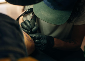 Close up of professional tattooer artist doing picture on hand of man by machine black ink from a jar.