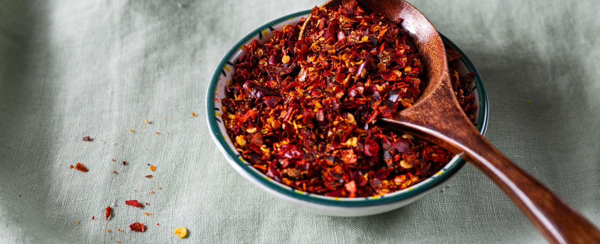 Bowl of crushed red pepper flakes with a wooden spoon on green linen fabric