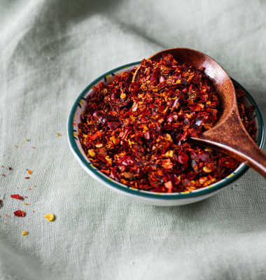 Bowl of crushed red pepper flakes with a wooden spoon on green linen fabric