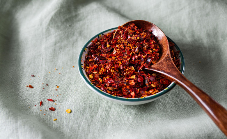 Bowl of crushed red pepper flakes with a wooden spoon on green linen fabric