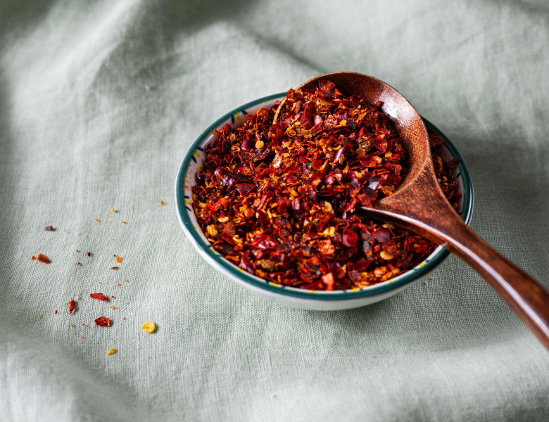 Bowl of crushed red pepper flakes with a wooden spoon on green linen fabric