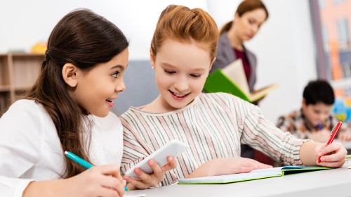 Smiling,Schoolgirls,Sitting,At,Desk,And,Using,Smartphone,During,Lesson