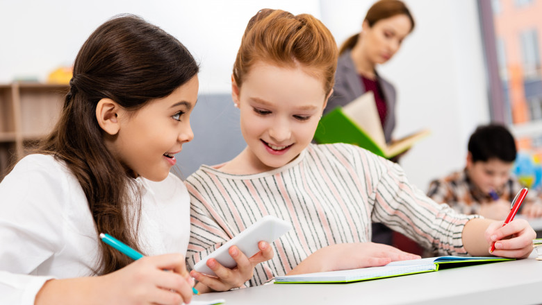 Smiling,Schoolgirls,Sitting,At,Desk,And,Using,Smartphone,During,Lesson