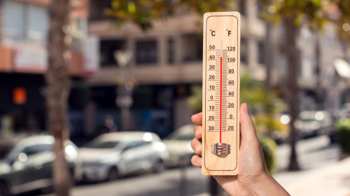 Hot,Weather.,Thermometer,In,Hand,In,Front,Of,An,Urban