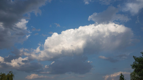 Beautiful cumulus clouds in blue sky with dark clouds on the edges and green trees below. Summer weather background for nature, relaxation, and enviro