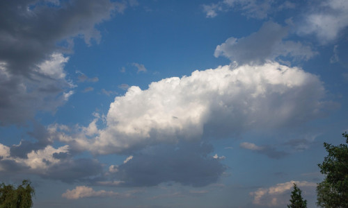 Beautiful cumulus clouds in blue sky with dark clouds on the edges and green trees below. Summer weather background for nature, relaxation, and enviro