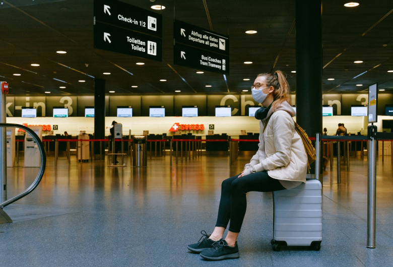 Femeie stă pe bagaj cu masca sanitară pe față, călătorie, turism în vremea COVID-19, coronavirus, aeroport