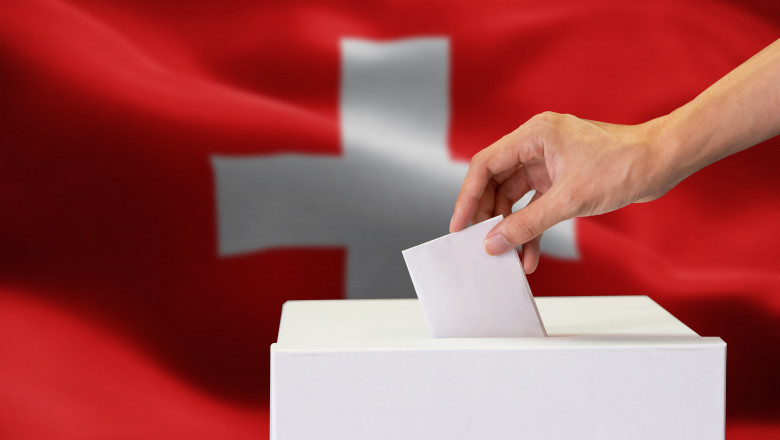 Close-up of human hand casting and inserting a vote and choosing and making a decision what he wants in polling box with Switzerland flag blended in background.