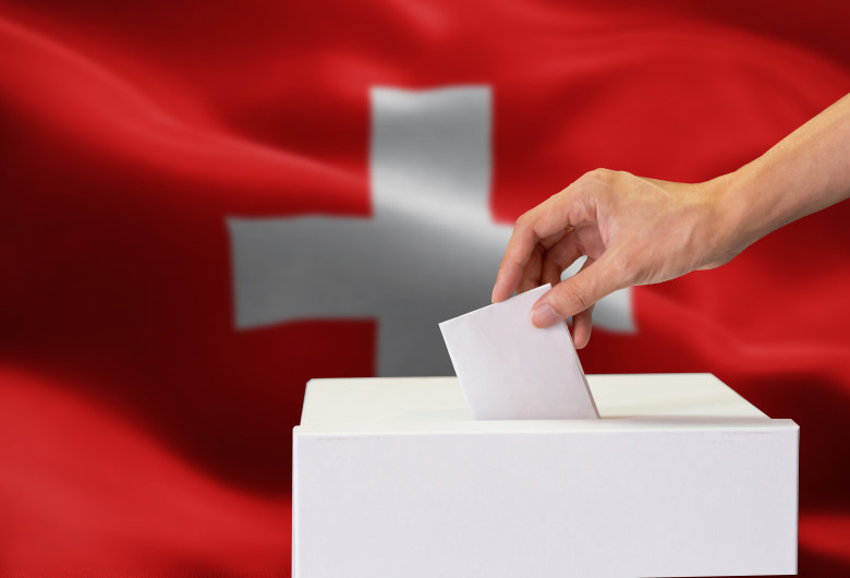 Close-up of human hand casting and inserting a vote and choosing and making a decision what he wants in polling box with Switzerland flag blended in background.