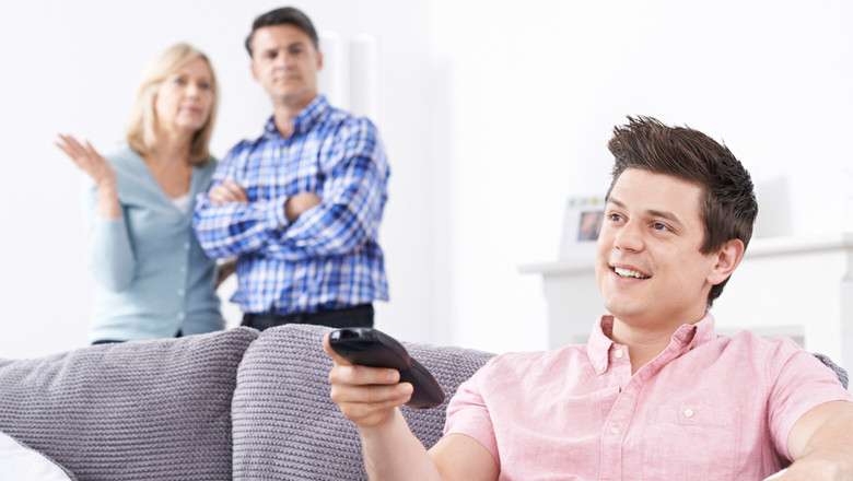 Tânăr stă acasă, în locuință cu părinții, se uită la televizor, mamă și tată supărați, frustrați de adolescent la TV