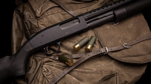 A black 12-gauge pump-action shotgun and ammo are on a canvas backpack. A smooth-bore weapon with a plastic stock. Dark background.