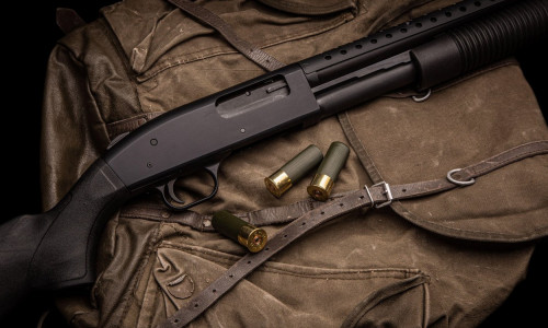 A black 12-gauge pump-action shotgun and ammo are on a canvas backpack. A smooth-bore weapon with a plastic stock. Dark background.