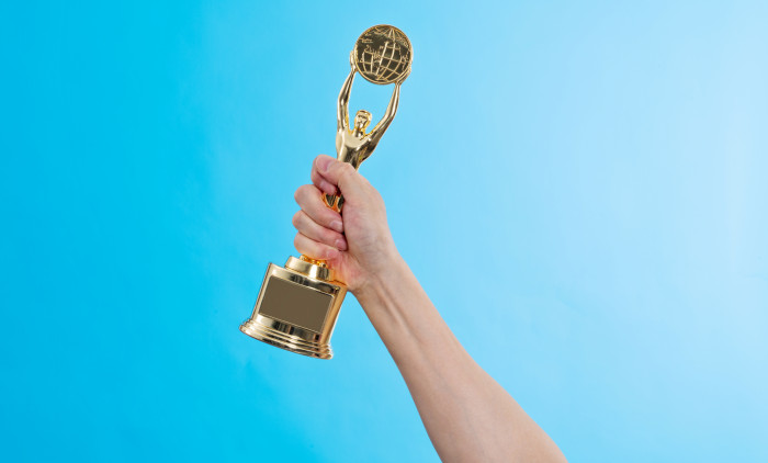 Hand,Holding,Golden,Statuette,Trophy,On,Blue,Background.