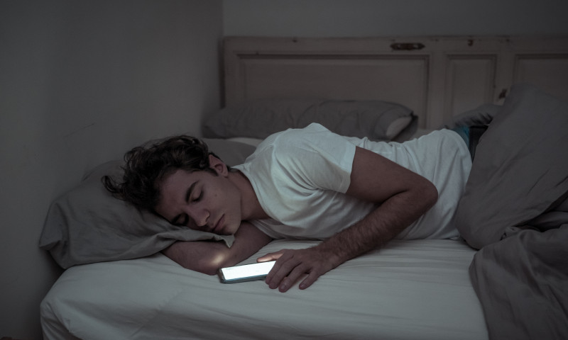 Addicted to social media young man falling asleep with smart mobile phone at night in bed. lifestyle portrait of man sleeping in dark bedroom with mobile screen light on. Mobile use addiction.