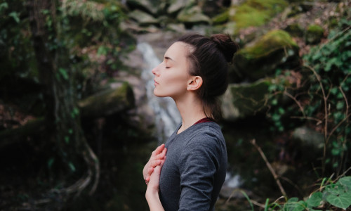 Young woman practicing breathing yoga pranayama outdoors in moss forest on background of waterfall. Unity with nature concept.